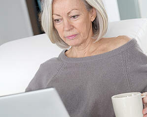 Ældre kvinde sidder alvorlig og ser på sin computer med en kop kaffe i hånden